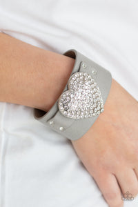 Flauntable Flirt - Silver - Paparazzi Accessories -   Item #P9DI-URSV-133XX Encrusted in blinding white rhinestones, an oversized silver heart frame is studded in place across the front of a gray leather band, creating a flirtatious centerpiece around the wrist. Features an adjustable snap closure.  Sold as one individual bracelet.