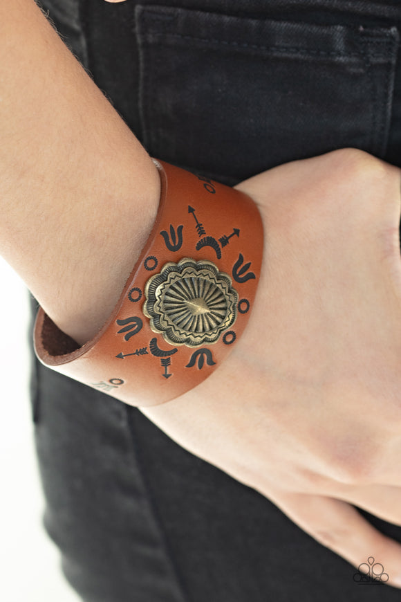 Desert Badlands - Brass/Brown - Paparazzi Accessories  An ornate brass frame is pressed into the center of a rust brown leather band stamped in desert inspired glyphs for a seasonal flair. Features an adjustable snap closure.  Sold as one individual.